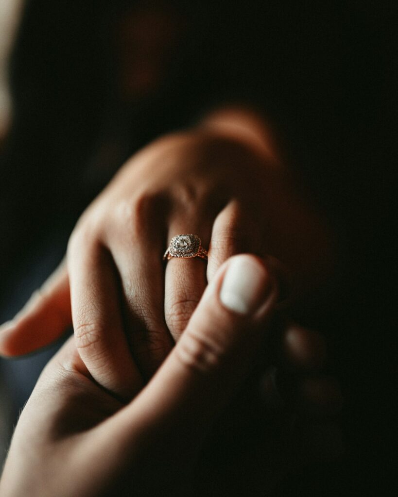 A hand wearing a diamond engagement ring, held gently in another hand